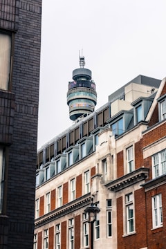 A cityscape featuring a tall telecommunications tower rising above a mix of modern and historic brick buildings. The architecture displays a combination of glass, steel, and traditional bricks with detailed window frames. The tower has multiple circular viewing platforms and antennas on top.