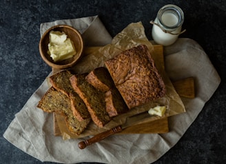 A rustic setup featuring a loaf of freshly baked bread, partly sliced and placed on parchment paper. A small knife with a wooden handle rests beside the slices, next to a serving of butter. A bowl containing more butter and a bottle of milk are also present. The background consists of dark textured surfaces and a natural fabric, giving a homey and warm feel.