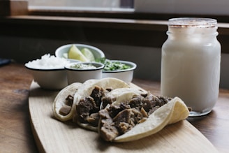 A rustic wooden table set with smoked meats, fresh tortillas, and colorful side dishes under warm natural light.