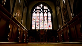 The image displays the interior of a church with large stained glass windows at the center. The windows are intricately designed with colorful depictions, casting a colorful glow inside. The wooden pews line both sides leading up to a central altar area. The architecture features high ceilings and detailed carvings.