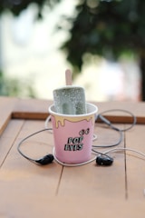 A popsicle is placed upright in a pink paper cup labeled 'POP EYES' with a playful design featuring cartoon eyes. The popsicle is green and has a frosty texture. Surrounding the cup is a pair of black earphones, and everything rests on a wooden surface, creating a casual and laid-back atmosphere.