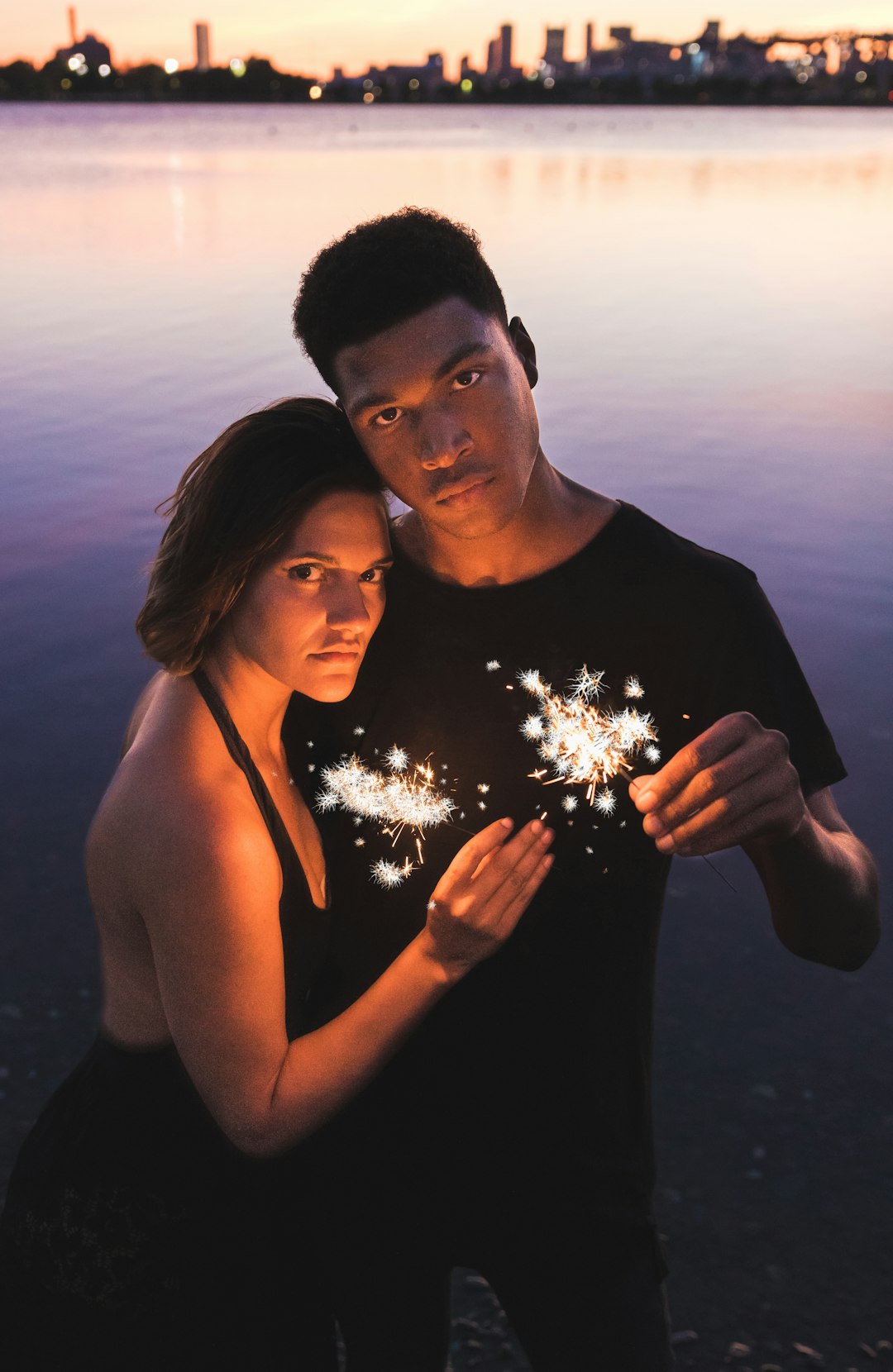 couple holding sparklers standing near body of water at dusk