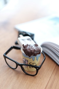 A chocolate-covered ice cream bar rests inside a small cup decorated with playful illustrations and the word 'CO' visible on it. Black-rimmed glasses frame the cup on a wooden surface, while an open book is slightly out of focus in the background.