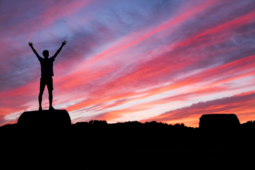 A silhouette of a man lifting up his hands while while standing on a rock at sunrise