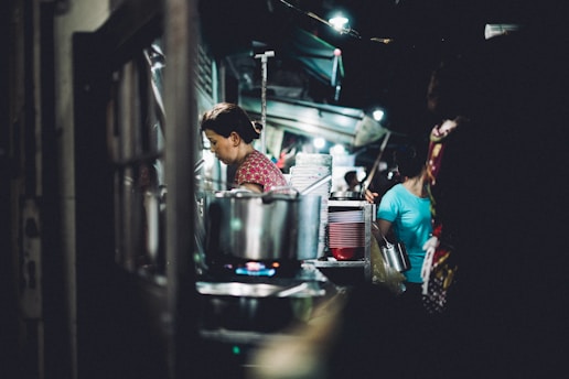 A woman is working in a dimly-lit street food stall. She is focused on her task, surrounded by cooking pots and a stack of plates. Another person is seen in the background wearing a blue shirt carrying a container, while the atmosphere suggests a busy market.