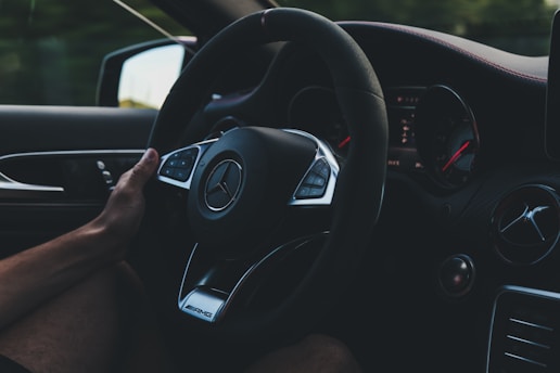 A hand is gripping the steering wheel of a luxury car, featuring the Mercedes-Benz logo prominently in the center. The dashboard and instrument panel are visible, with the RPM and speed gauges slightly illuminated against a dark background.