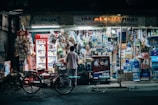 A small, bustling convenience store with a bright, neon-lit exterior. Various items including beverages, snacks, and household goods are neatly displayed. A red refrigerator with Coca-Cola branding is prominent, and a bicycle is parked in front of the store. Two people are interacting at the counter, suggesting a friendly, community atmosphere.