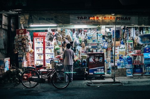 A small, bustling convenience store with a bright, neon-lit exterior. Various items including beverages, snacks, and household goods are neatly displayed. A red refrigerator with Coca-Cola branding is prominent, and a bicycle is parked in front of the store. Two people are interacting at the counter, suggesting a friendly, community atmosphere.