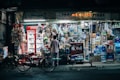 A small, bustling convenience store with a bright, neon-lit exterior. Various items including beverages, snacks, and household goods are neatly displayed. A red refrigerator with Coca-Cola branding is prominent, and a bicycle is parked in front of the store. Two people are interacting at the counter, suggesting a friendly, community atmosphere.