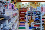 A colorful and cluttered candy store with a variety of sweets and snacks displayed on shelves and in dispensers. The left side features an array of candy dispensers, while the right has shelves stocked with boxes and packets of snacks. The back wall has a display of Pez dispensers and hanging decorations. Balloons and various signage add to the festive atmosphere.
