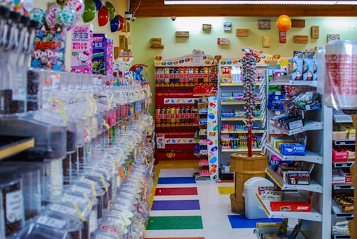 A colorful and cluttered candy store with a variety of sweets and snacks displayed on shelves and in dispensers. The left side features an array of candy dispensers, while the right has shelves stocked with boxes and packets of snacks. The back wall has a display of Pez dispensers and hanging decorations. Balloons and various signage add to the festive atmosphere.