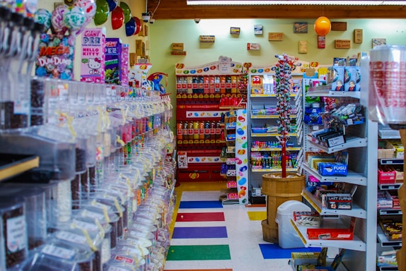 A colorful and cluttered candy store with a variety of sweets and snacks displayed on shelves and in dispensers. The left side features an array of candy dispensers, while the right has shelves stocked with boxes and packets of snacks. The back wall has a display of Pez dispensers and hanging decorations. Balloons and various signage add to the festive atmosphere.