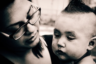 A black and white close-up photograph featuring a woman with glasses holding a baby. The woman's face is tilted downward with a gentle, caring expression. The baby has a somber look with visible textures on the skin and a distinct hairstyle.