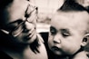 A black and white close-up photograph featuring a woman with glasses holding a baby. The woman's face is tilted downward with a gentle, caring expression. The baby has a somber look with visible textures on the skin and a distinct hairstyle.