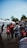 Several people are gathered outdoors in what appears to be an area of tents and trailers. Some individuals are walking or standing, and others are engaged in conversation. One person is sweeping the ground while another person is on the phone. There is a mix of adults and children, and some people are wearing backpacks or carrying items. The scene has a casual and lively atmosphere with trees visible in the background.