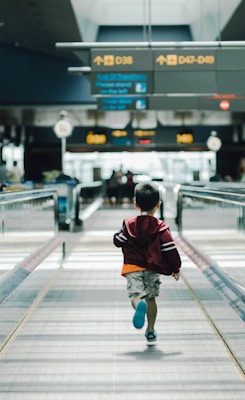 A young child wearing a maroon hoodie and shorts is walking along a moving walkway in an airport. Overhead signs indicate directions to gates, and the background is slightly blurred, suggesting movement.