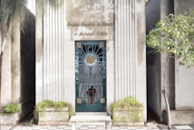 A decorative metal door set between two fluted stone columns with engraved text at the top. The door features intricate metalwork with circular and geometric patterns, positioned on a small stepped platform with tiled flooring. Surrounding the entrance are small planters with green foliage and a tree on the right side.