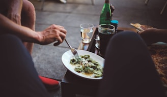 A person is enjoying a meal outdoors. A plate of ravioli with sauce is on a small table. Nearby, there's a glass with a dark beverage, a wine glass with a light drink, and a green bottle. The setting appears casual and relaxed, suggesting a dining experience. Another person's hand is reaching for a piece of pizza.