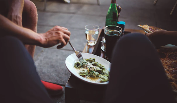 A person is enjoying a meal outdoors. A plate of ravioli with sauce is on a small table. Nearby, there's a glass with a dark beverage, a wine glass with a light drink, and a green bottle. The setting appears casual and relaxed, suggesting a dining experience. Another person's hand is reaching for a piece of pizza.