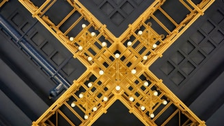 An intricate ceiling design featuring a symmetrical arrangement of golden geometric patterns intersecting to form an elaborate cross-like structure. The ceiling is embellished with circular white lights, adding contrast to the dark blue background grid pattern of the ceiling.