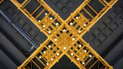 An intricate ceiling design featuring a symmetrical arrangement of golden geometric patterns intersecting to form an elaborate cross-like structure. The ceiling is embellished with circular white lights, adding contrast to the dark blue background grid pattern of the ceiling.
