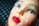 A realistic-looking mannequin head with vivid red lips and detailed facial features is encased in a layer of clear plastic wrap. The background features a textured dark gray grid pattern.