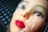 A realistic-looking mannequin head with vivid red lips and detailed facial features is encased in a layer of clear plastic wrap. The background features a textured dark gray grid pattern.