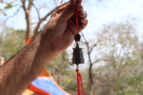 A hand holding a small pagoda-shaped ornament with red tassels against a blurry natural background of trees.
