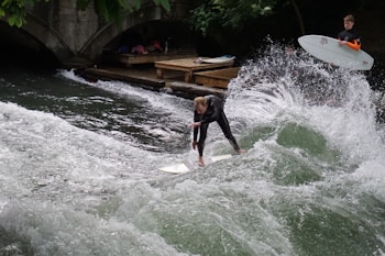 A person in a wetsuit surfs on a standing wave in a river while another person stands on a wooden platform nearby holding a surfboard. The water splashes around the surfer, creating dynamic movement and energy. A stone bridge arches in the background.