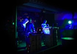 Two individuals are performing music in a dimly lit venue, surrounded by DJ equipment and laptops. The walls are decorated with graffiti, and colorful LED lights cast a blue-green hue over the scene, creating a vibrant atmosphere.