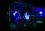 Two individuals are performing music in a dimly lit venue, surrounded by DJ equipment and laptops. The walls are decorated with graffiti, and colorful LED lights cast a blue-green hue over the scene, creating a vibrant atmosphere.