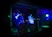Two individuals are performing music in a dimly lit venue, surrounded by DJ equipment and laptops. The walls are decorated with graffiti, and colorful LED lights cast a blue-green hue over the scene, creating a vibrant atmosphere.