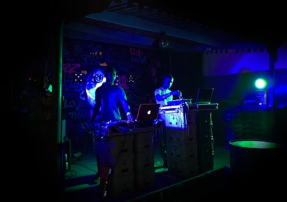 Two individuals are performing music in a dimly lit venue, surrounded by DJ equipment and laptops. The walls are decorated with graffiti, and colorful LED lights cast a blue-green hue over the scene, creating a vibrant atmosphere.