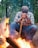 A person sitting on a log in a forest with their head resting on a stick, appearing contemplative. In the foreground, a campfire is burning brightly, providing warmth and light. The surrounding area is filled with tall trees, indicating a woodland setting.