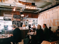 A cozy cafe interior with wooden accents and industrial lighting. Various patrons are seated at round tables, engaged in conversation or on their devices. The wall features the words 'BANG BANG' in large letters. A menu board with various offerings like coffee and breakfast is prominently displayed.