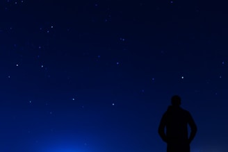 A silhouette of a person stands under a clear night sky filled with numerous bright stars. The scene conveys a sense of solitude and wonder as the silhouette gazes upwards.