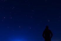A silhouette of a person stands under a clear night sky filled with numerous bright stars. The scene conveys a sense of solitude and wonder as the silhouette gazes upwards.