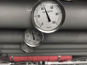Several industrial thermometers are mounted on large metallic pipes. The environment has a utilitarian feel, consistent with a mechanical or industrial setting.