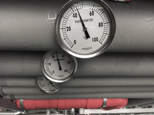 Several industrial thermometers are mounted on large metallic pipes. The environment has a utilitarian feel, consistent with a mechanical or industrial setting.