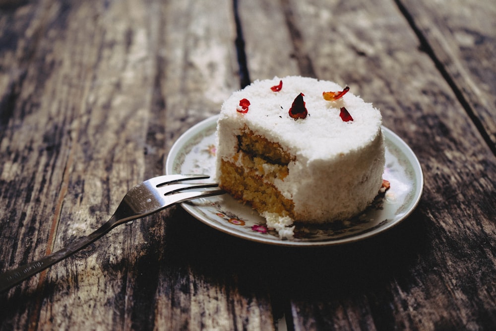 round icing-covered white cake on round white and green floral ceramic saucer beside gray fork on brown wooden table