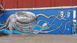 A vibrant mural depicting a large, detailed octopus with long tentacles extending across a wall. The octopus is rendered in black and white, while the background is a bright, deep blue. The tentacles interact with different theatrical masks and objects, including a skull and additional masks. A sign reading 'THEATRE' is visible, along with a character dressed in theatrical costume.