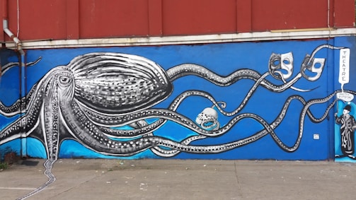 A vibrant mural depicting a large, detailed octopus with long tentacles extending across a wall. The octopus is rendered in black and white, while the background is a bright, deep blue. The tentacles interact with different theatrical masks and objects, including a skull and additional masks. A sign reading 'THEATRE' is visible, along with a character dressed in theatrical costume.