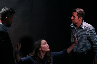 A dimly lit scene featuring three people engaged in a tense interaction. A woman with long hair appears to be pleading or explaining something while gesturing with her hands. Two men stand on either side, both with serious expressions, as they listen intently to the woman.