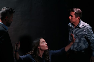A dimly lit scene featuring three people engaged in a tense interaction. A woman with long hair appears to be pleading or explaining something while gesturing with her hands. Two men stand on either side, both with serious expressions, as they listen intently to the woman.