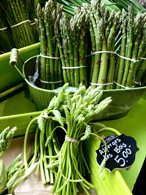 A collection of fresh green asparagus stalks neatly bundled and displayed in a market setting. A small chalkboard sign indicates the price of the asparagus in euros.