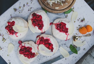 A top-down view of several meringue desserts topped with whipped cream and fresh raspberries, placed on a white marble surface. Sprinkled flower petals and small green leaves add a decorative touch around the desserts. An ornate spoon and two small orange fruits are positioned nearby. A wooden board with shredded cheese or chocolate sits in the upper part of the composition.