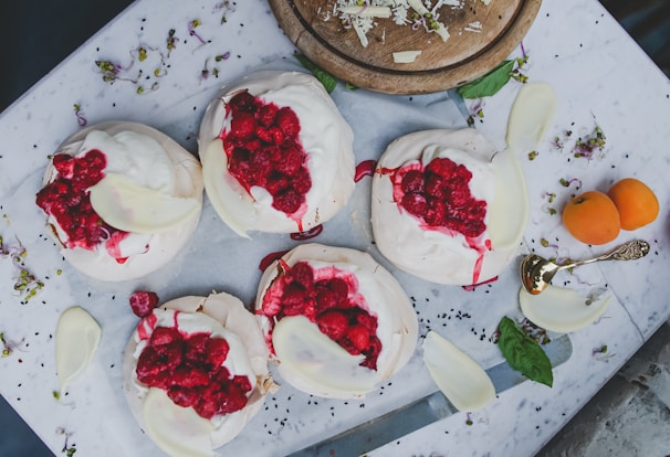A top-down view of several meringue desserts topped with whipped cream and fresh raspberries, placed on a white marble surface. Sprinkled flower petals and small green leaves add a decorative touch around the desserts. An ornate spoon and two small orange fruits are positioned nearby. A wooden board with shredded cheese or chocolate sits in the upper part of the composition.