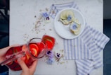 A person is pouring a bright red drink, containing slices of citrus and cucumber, from a pitcher into a glass. The setting includes a white marble table with a blue and white striped cloth. On the table, there is a white plate with two pieces of bread topped with slices of green pear and some decorative blue flowers. Additional purple flowers and some food crumbs are scattered around the table.