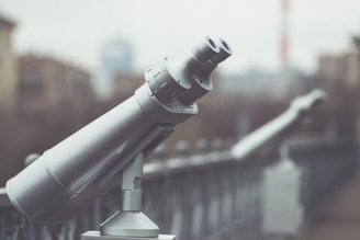 A row of public observation binoculars is mounted along a railing, with a blurred urban backdrop. The foreground binoculars are gray and metallic, standing out against the softer, muted background.