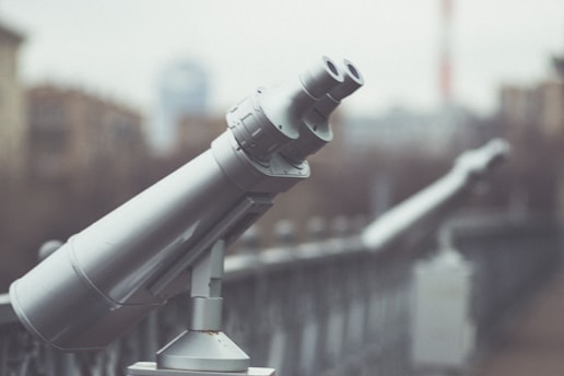A row of public observation binoculars is mounted along a railing, with a blurred urban backdrop. The foreground binoculars are gray and metallic, standing out against the softer, muted background.